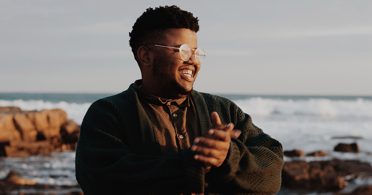 Young man with glasses, rubbing his hands together with the sea and cliffs in the background.