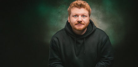 A man with short reddish-blonde hair and a beard wearing a black hoodie looks slightly upward. The background is a textured mix of green and brown tones.