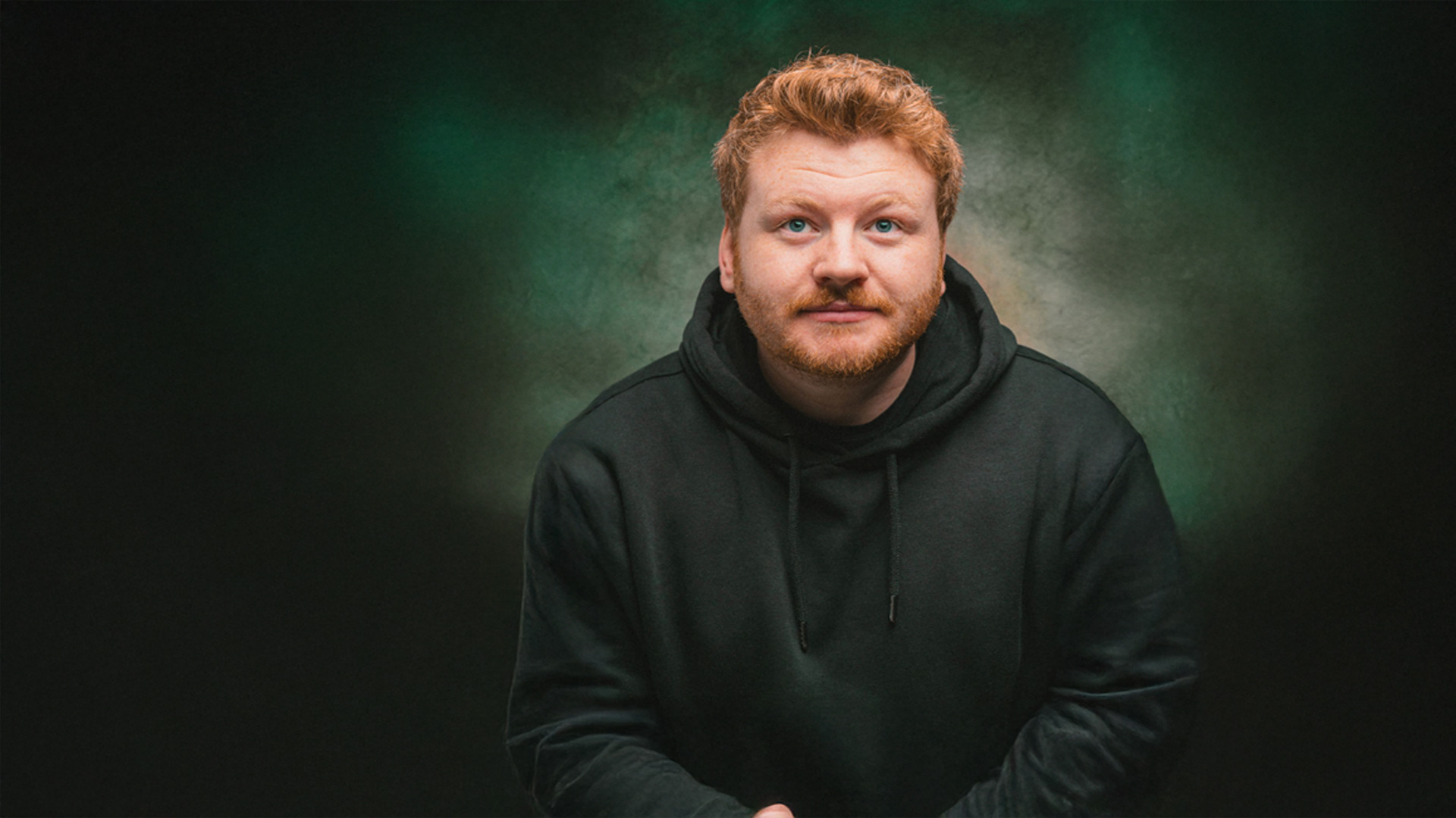 A man with short reddish-blonde hair and a beard wearing a black hoodie looks slightly upward. The background is a textured mix of green and brown tones.
