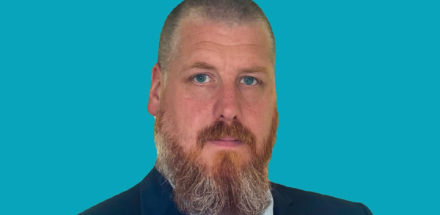 A man with short hair and a full reddish-grey beard wearing a dark suit, white shirt, and tie, posing against a solid turquoise background.