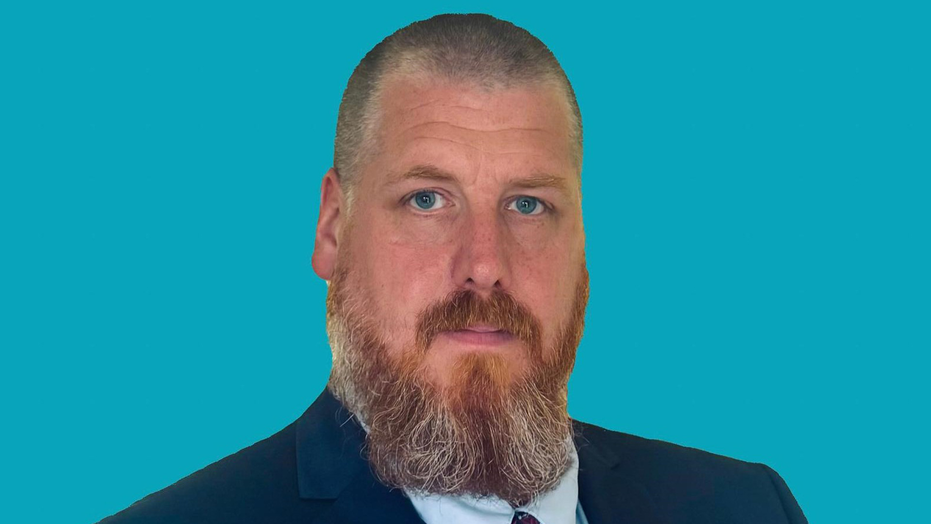 A man with short hair and a full reddish-grey beard wearing a dark suit, white shirt, and tie, posing against a solid turquoise background.
