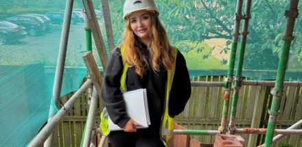 A young woman wearing a hard hat and high-visibility vest stands on scaffolding at a construction site.